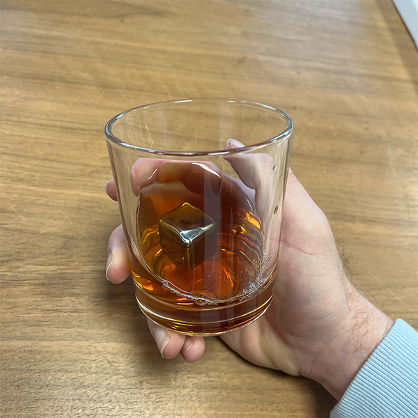 plain_whiskey_tumbler_with_whiskey_inside_and_stainless_steel_cooling_stone_in_hand